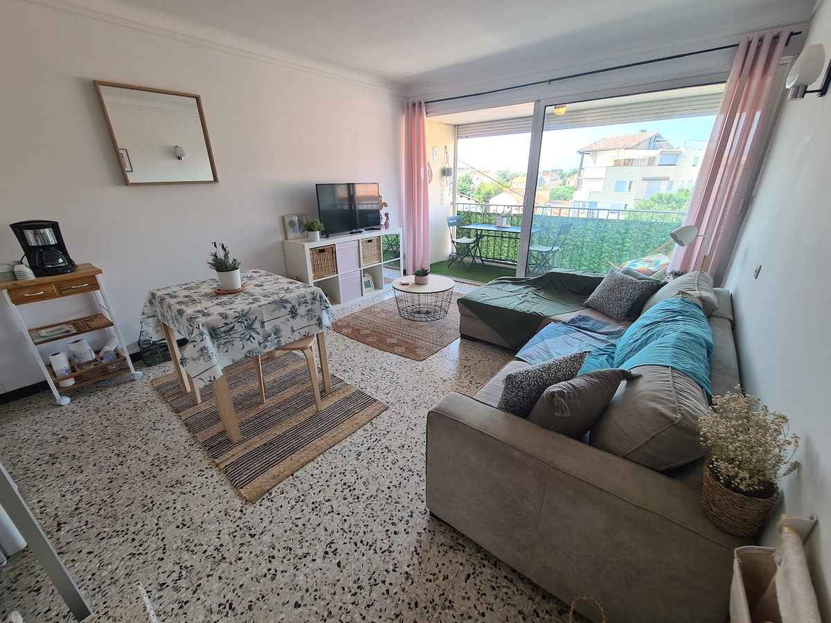 A well-furnished living area features a light-colored sectional sofa and a coffee table. A dining table with floral decor is positioned near a large window. Natural light fills the space, highlighting a television and a small side table. A balcony is visible through the sliding glass doors.
