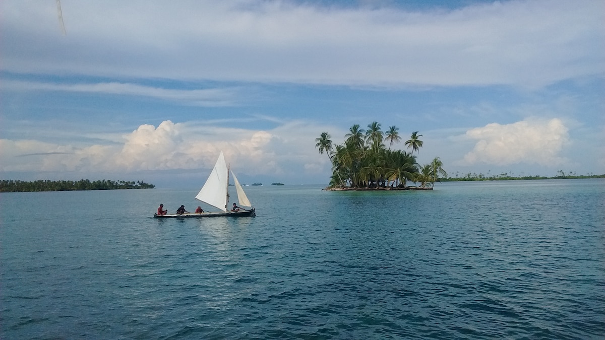 A sailboat with a white sail is seen gliding across calm turquoise waters. In the background, a small island with palm trees stands against a clear sky, creating a serene and inviting scene typical of the San Blas Islands.