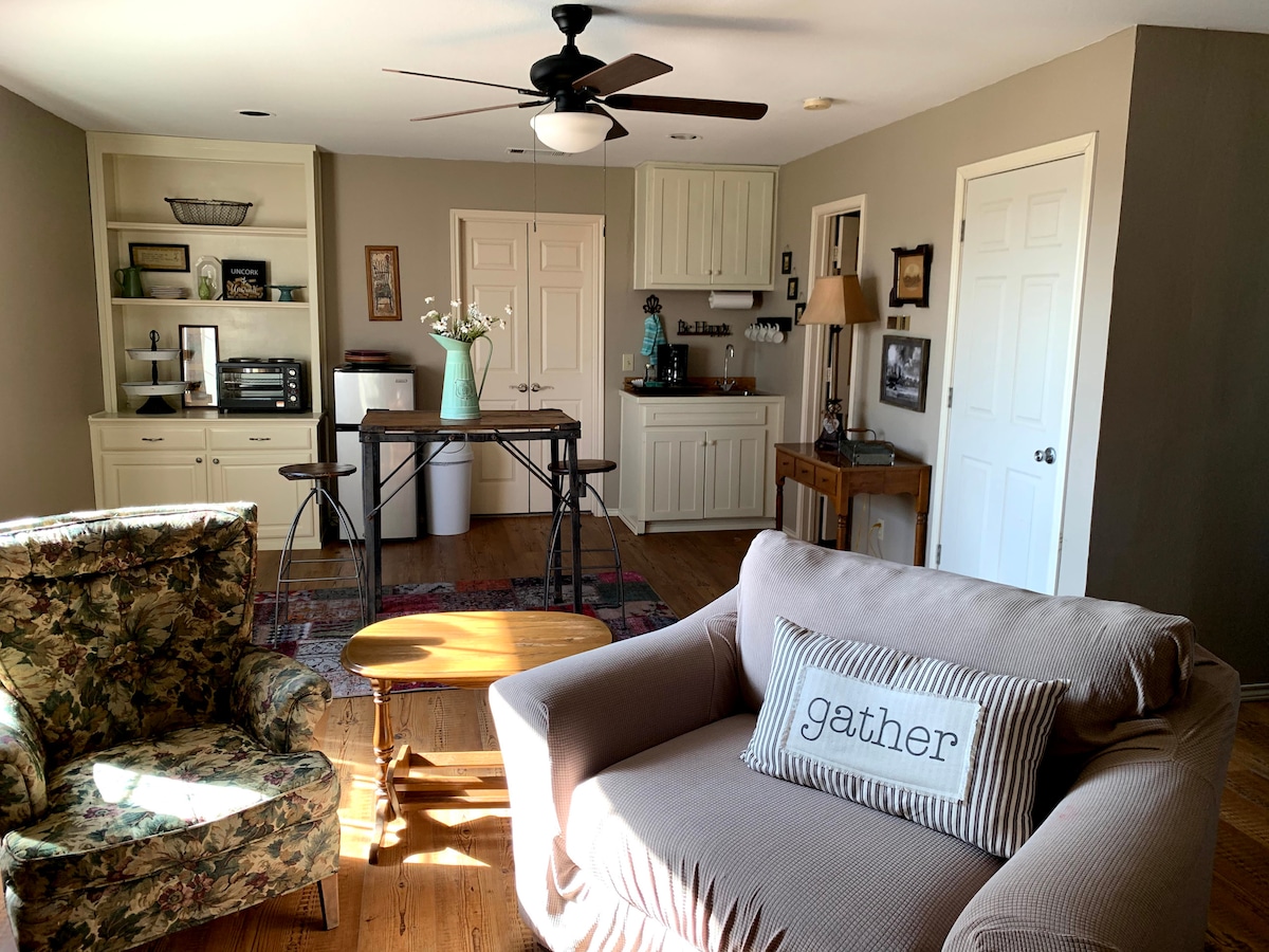 An inviting open living area features a comfortable sofa with a decorative pillow and an armchair with a floral pattern. A small wooden coffee table sits in front of the seating. The kitchenette is visible in the background, showing cream-colored cabinetry and a small dining table.