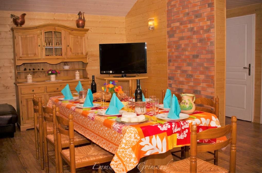 A dining area features a large table set for a meal, adorned with a colorful tablecloth and blue napkins. A wooden sideboard is positioned against the wall, complemented by a flat-screen television. Natural wood tones create a warm ambiance.