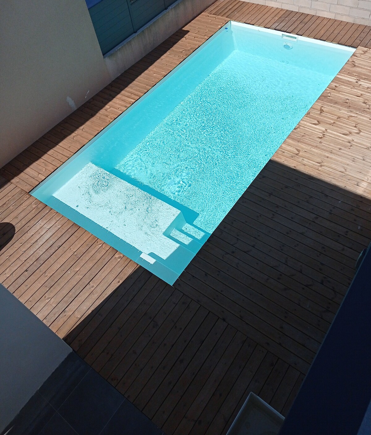 A modern rectangular pool is surrounded by wooden decking, providing an inviting space. The clear blue water reflects sunlight, contrasting with the surrounding walls. A section for entry steps can be seen at one end, enhancing accessibility to the pool area.