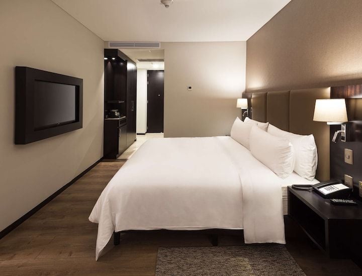 A modern bedroom features a spacious bed with crisp white linens. A flat-screen television is mounted on the wall opposite the bed. Soft lighting is provided by bedside lamps, and warm wooden flooring adds to the inviting ambiance.