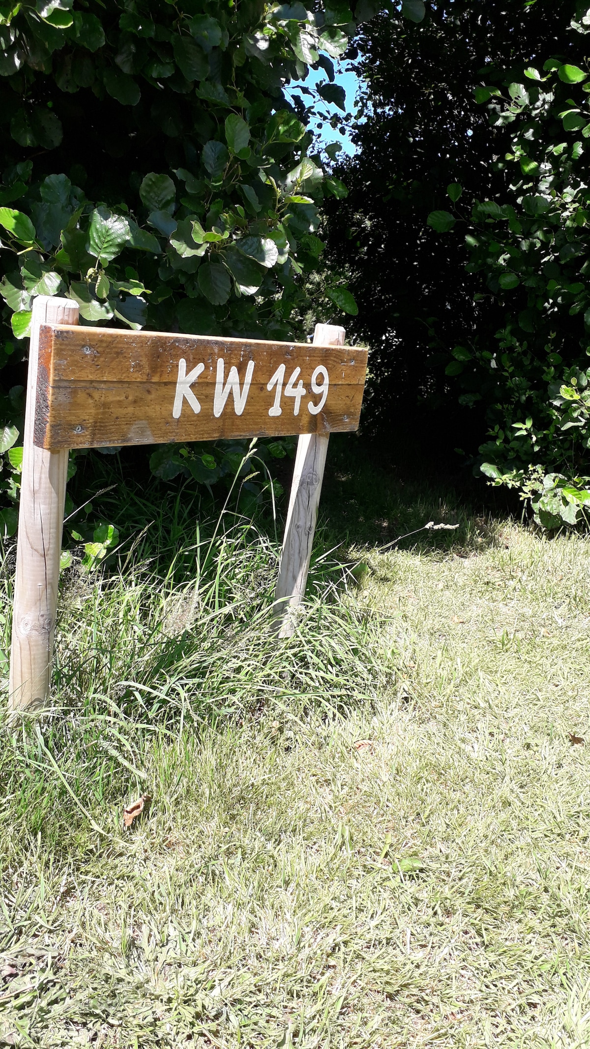 A wooden sign marked 'KW 149' is supported by two posts, surrounded by greenery. The grass is kept short, and the foliage creates a natural frame around the sign, highlighting its location within a peaceful outdoor area.
