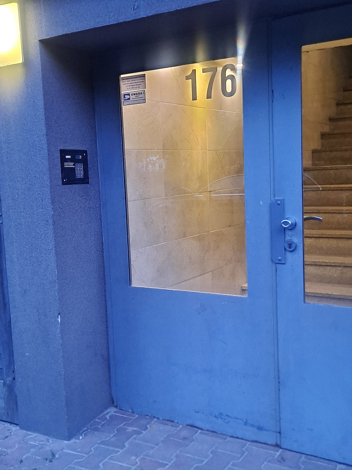 The entrance door is framed by a concrete wall, featuring large glass panels and the number '176' prominently displayed. A secure entry system is located to the left of the door, and a staircase is visible through the glass, leading upwards.