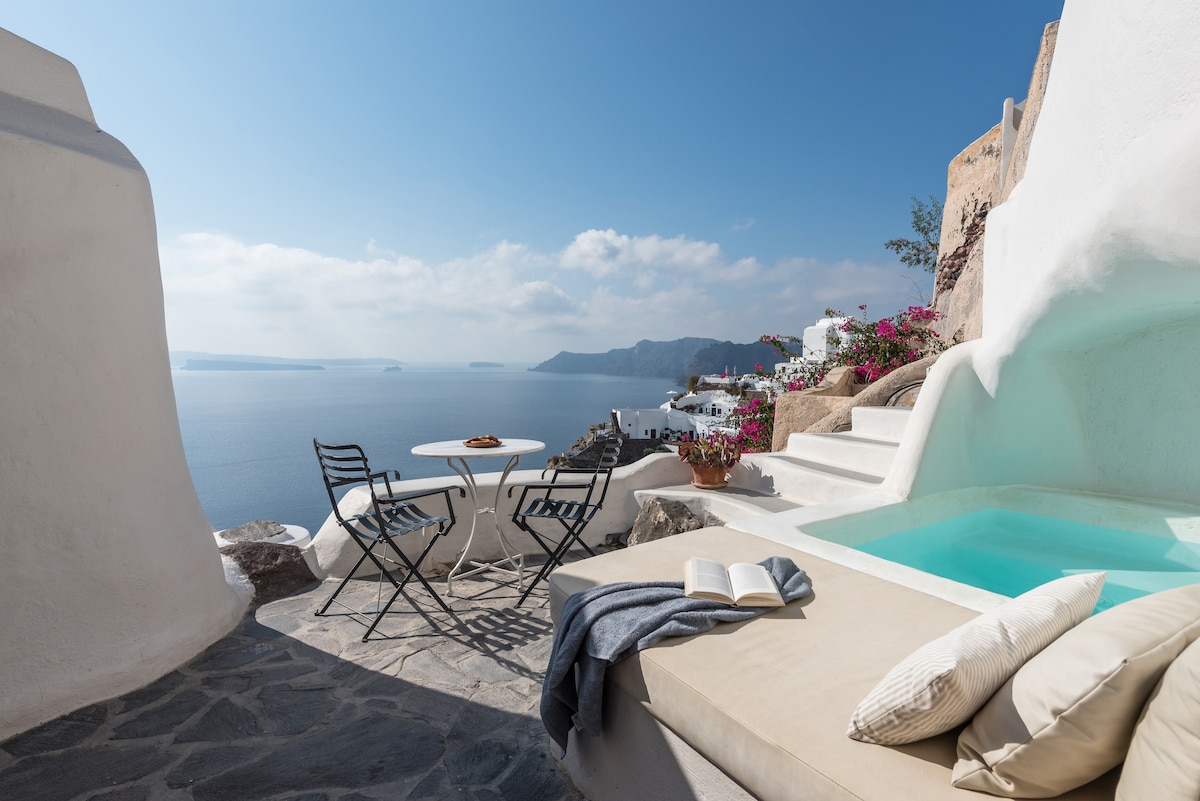 A private terrace is visible, featuring a light-colored seating area and a small round table with two chairs. A whirlpool is integrated into the space, creating a calm ambiance. The expansive view showcases the caldera and sea, complemented by vibrant flowers in the background.