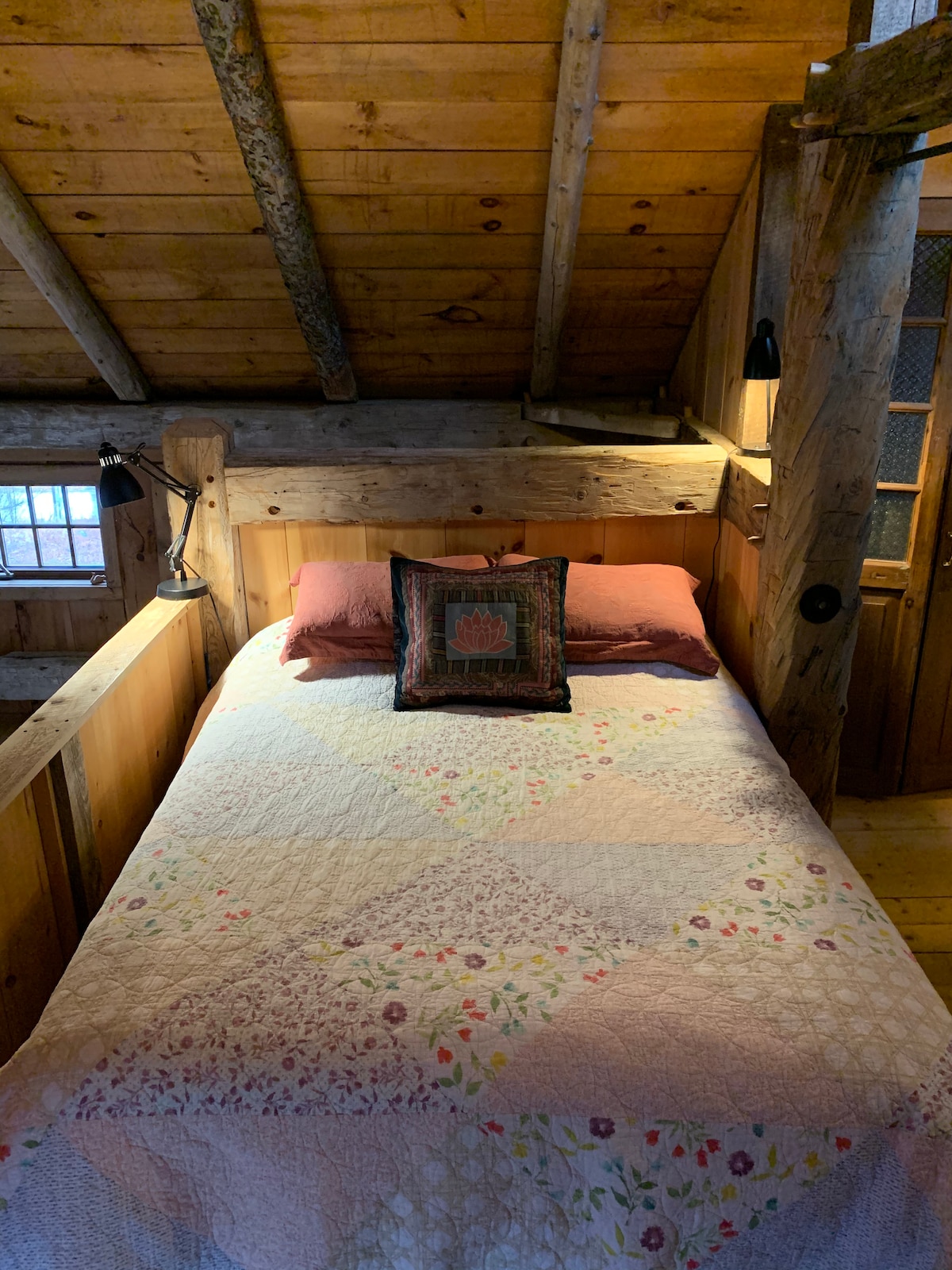 A cozy sleeping area features a spacious bed with a floral-patterned quilt. Accent pillows add subtle color. Soft lighting is provided by wall-mounted sconces. Exposed wooden beams and paneling contribute to a rustic, cabin-like ambiance, while a nearby wooden door hints at privacy.