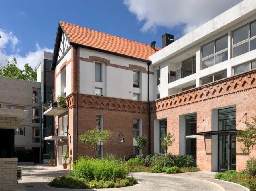 The building features a blend of modern and traditional architecture, showcasing brick and white stucco exteriors. Lush greenery and well-maintained landscaping complement the structure, while large windows allow natural light to illuminate the interiors.