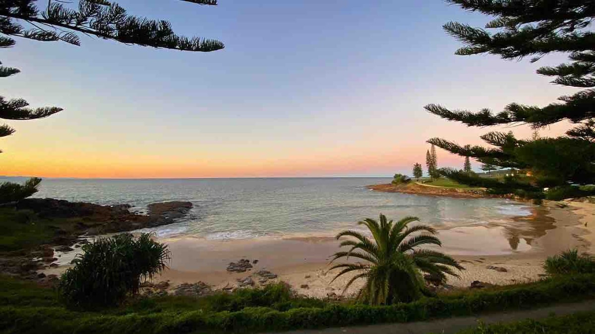 A serene beach scene is displayed, featuring gentle waves lapping at the shore. Palm trees frame the view, while the sky transitions from soft pink to warm orange hues as sunset approaches. The surrounding landscape includes rocky outcrops and lush greenery.