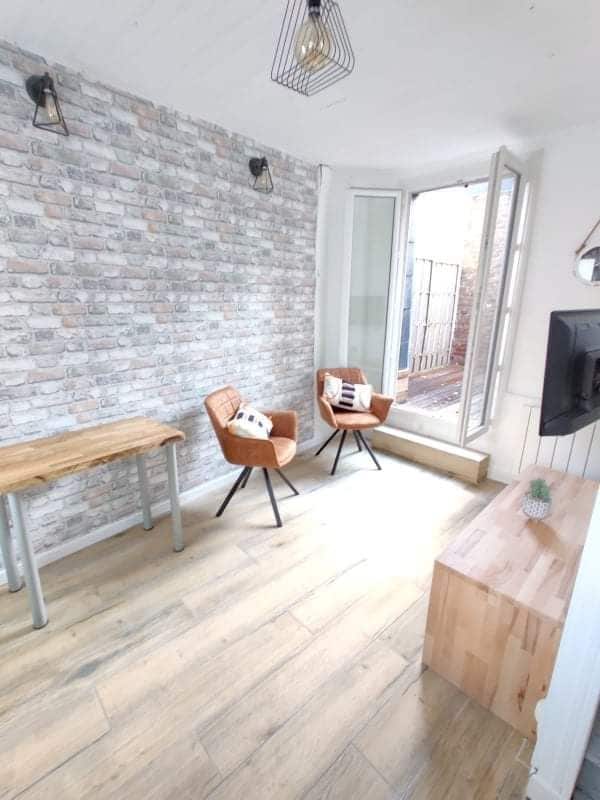 A small living area features two brown leather chairs positioned across from a wooden table and a light wood TV stand. Natural light enters through large open doors that lead to an outdoor space, while textured brick wallpaper adds a distinctive backdrop.