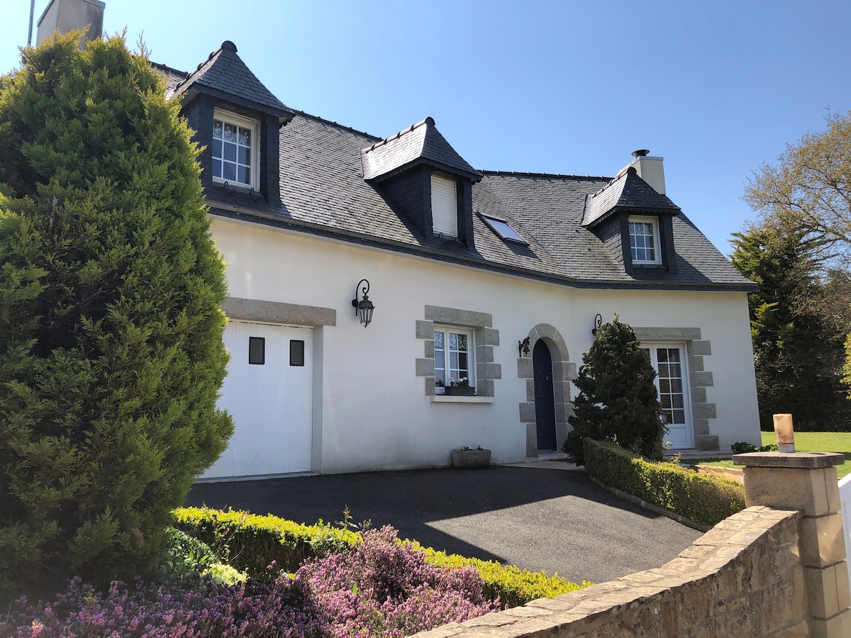 A charming neo-Breton family house is showcased, featuring a classic slate roof and distinctive white walls. The front garden is lush, with neatly trimmed shrubs and colorful flowerbeds. A driveway leads to an attached garage, while window shutters add a touch of character to the facade.