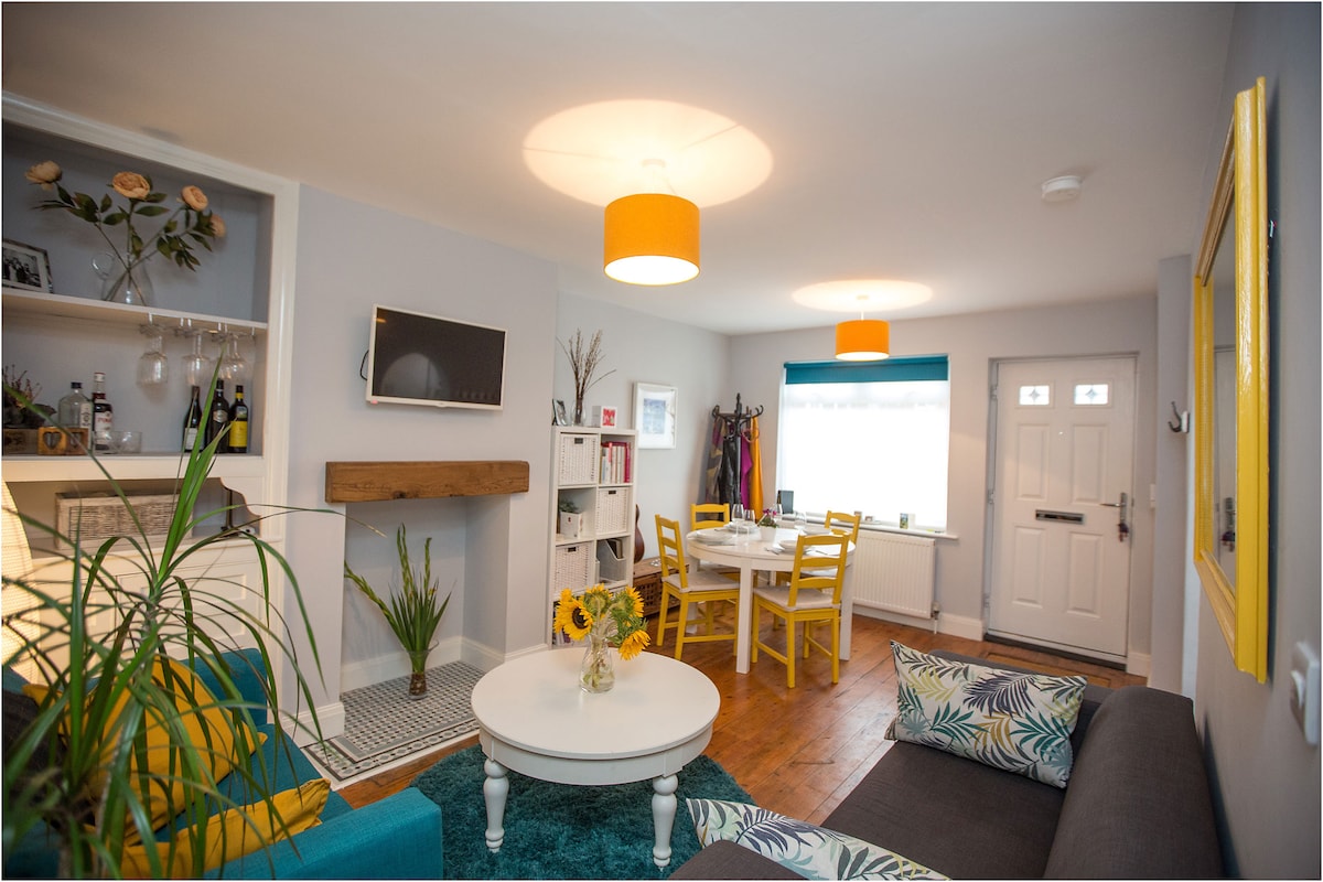 The lounge and dining area features a combination of bright yellow and turquoise decor. A comfortable sofa is positioned alongside a vibrant coffee table, with a potted plant nearby. Natural light enters through a window covered with a blue valance, illuminating the dining table set for four.