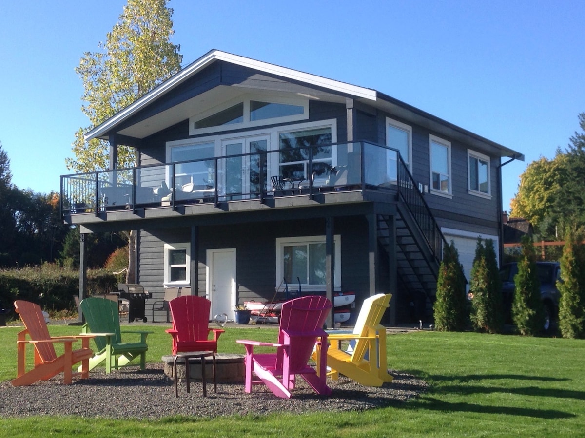 The two-story carriage house suite is showcased, featuring a spacious upper deck with glass railings. Colorful Adirondack chairs are arranged around a gravel area in front. Lush greenery and tidy landscaping frame the property, highlighting the inviting outdoor space.