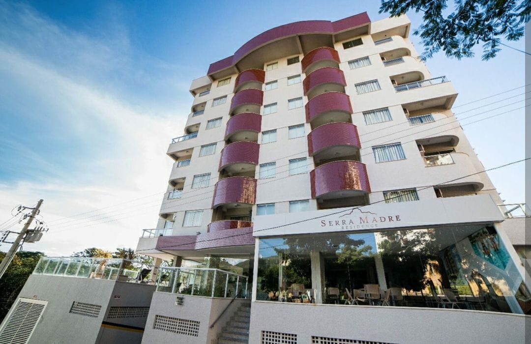A modern multi-story building stands under a clear blue sky, featuring a combination of soft colors and unique architectural details. The entrance area is accessible, with a glass front visible, inviting guests to explore the amenities offered within the condominium.