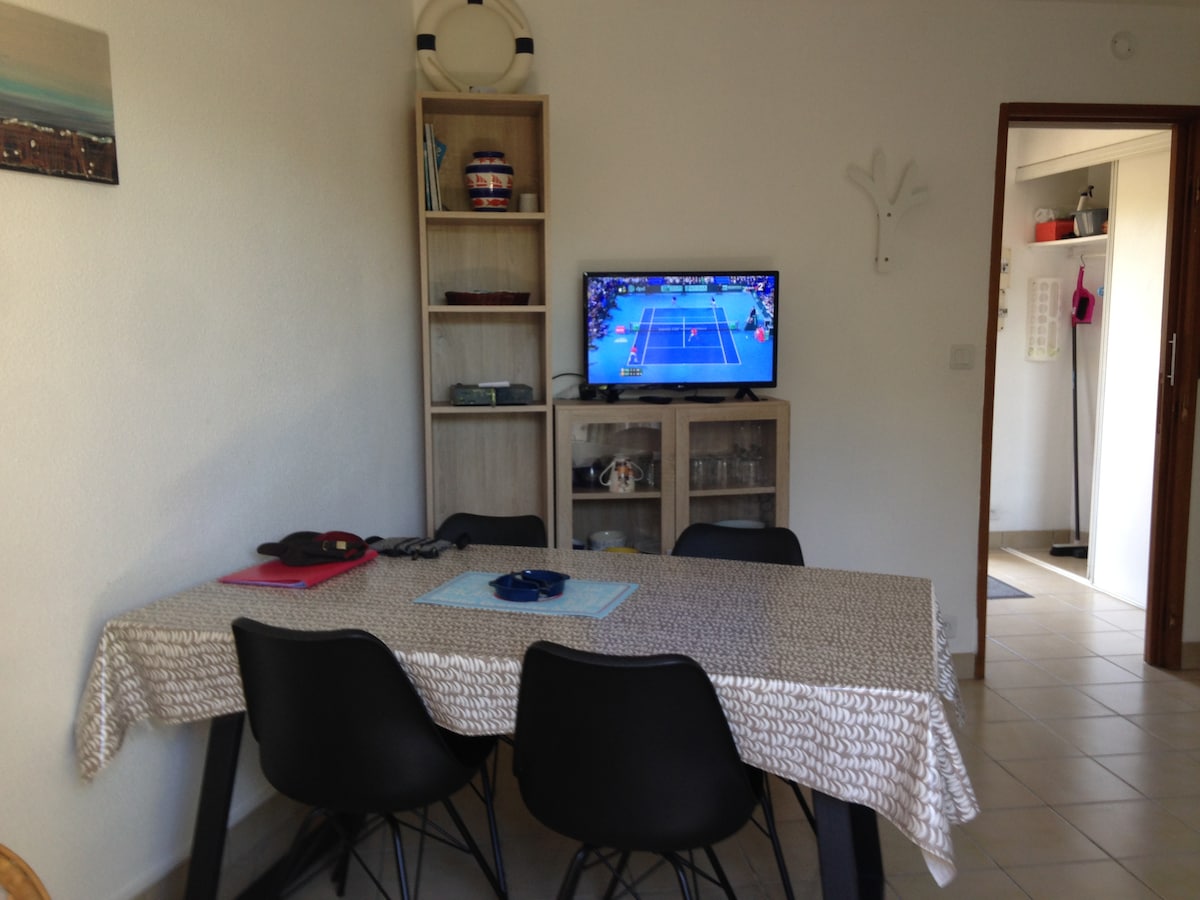 A dining area is presented with a rectangular table set with a tablecloth. Black chairs surround the table, creating a cozy dining space. A wall-mounted television displays a sports event, while a bookshelf with various decorative items is positioned in the corner.