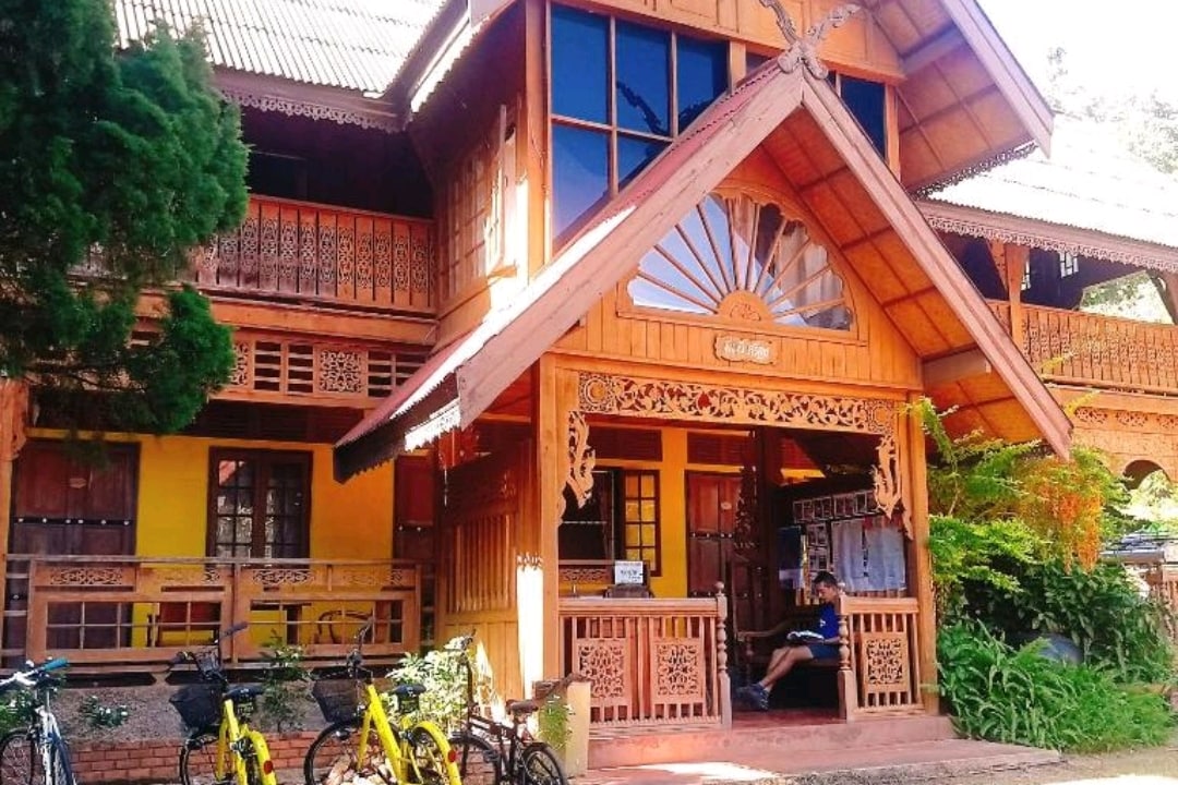 A charming two-story wooden house features intricate carvings and a welcoming entrance. Large windows allow natural light to fill the space, while bicycles are positioned near the front, offering a convenient option for exploration. Lush greenery surrounds the building, enhancing its inviting appearance.