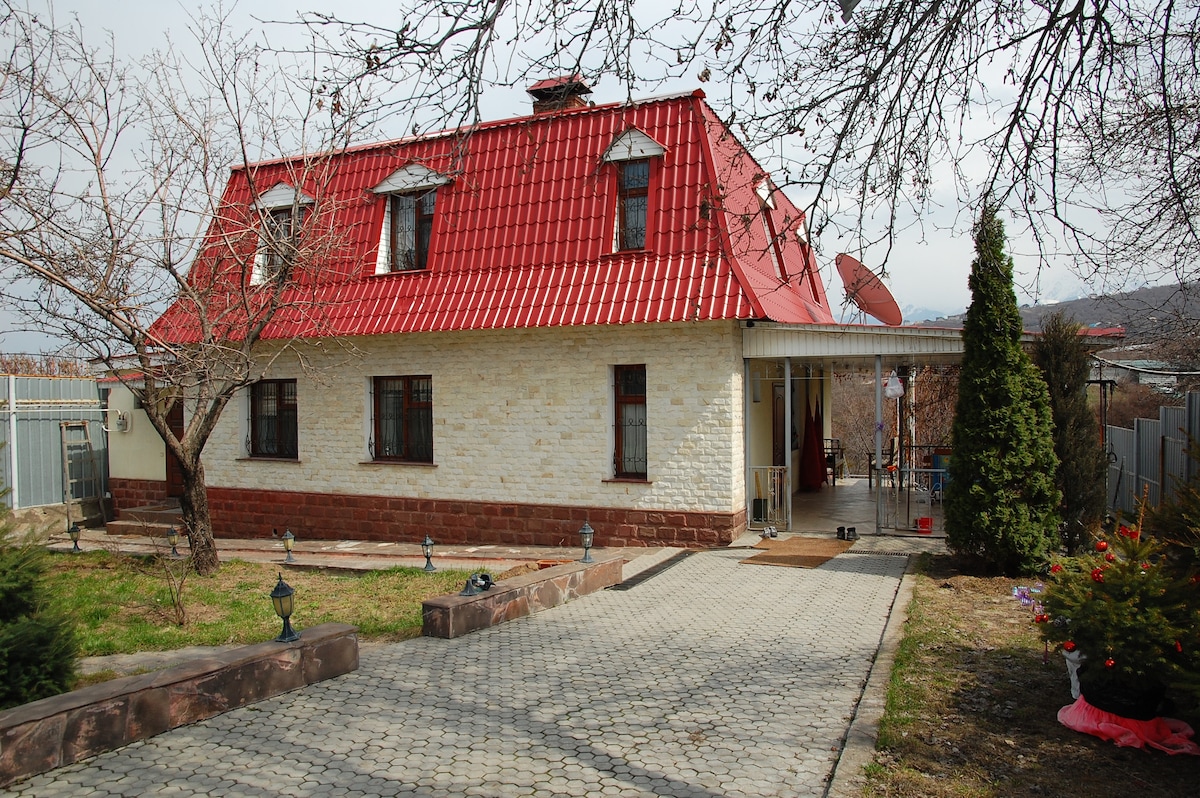 The exterior of the house features a distinctive red roof and a light stone façade. Symmetrical windows adorn the front, framed by a small garden area with trees and shrubs. A paved walkway leads to the entrance, providing a welcoming pathway.