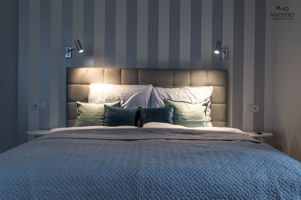 A spacious bed with a light gray headboard is positioned centrally against striped walls. Soft pillows in varying shades of green are arranged atop the bedding. Wall-mounted reading lamps provide focused lighting on either side, enhancing the serene sleeping environment.