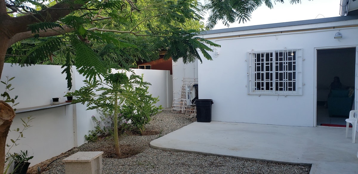 An outdoor area features a gravel surface bordered by green foliage and small trees. A white wall encloses the space, and a window with protective bars is visible on the building. A simple chair and a black trash bin are placed nearby, adding to the functional aesthetic.