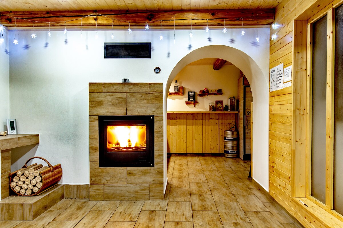 A warm and inviting common area features a modern wood-burning stove nestled in a stone facade, surrounded by wooden paneling. The space is enhanced by soft lighting, creating a cozy atmosphere. A corner displays neatly stacked firewood, ready for use.