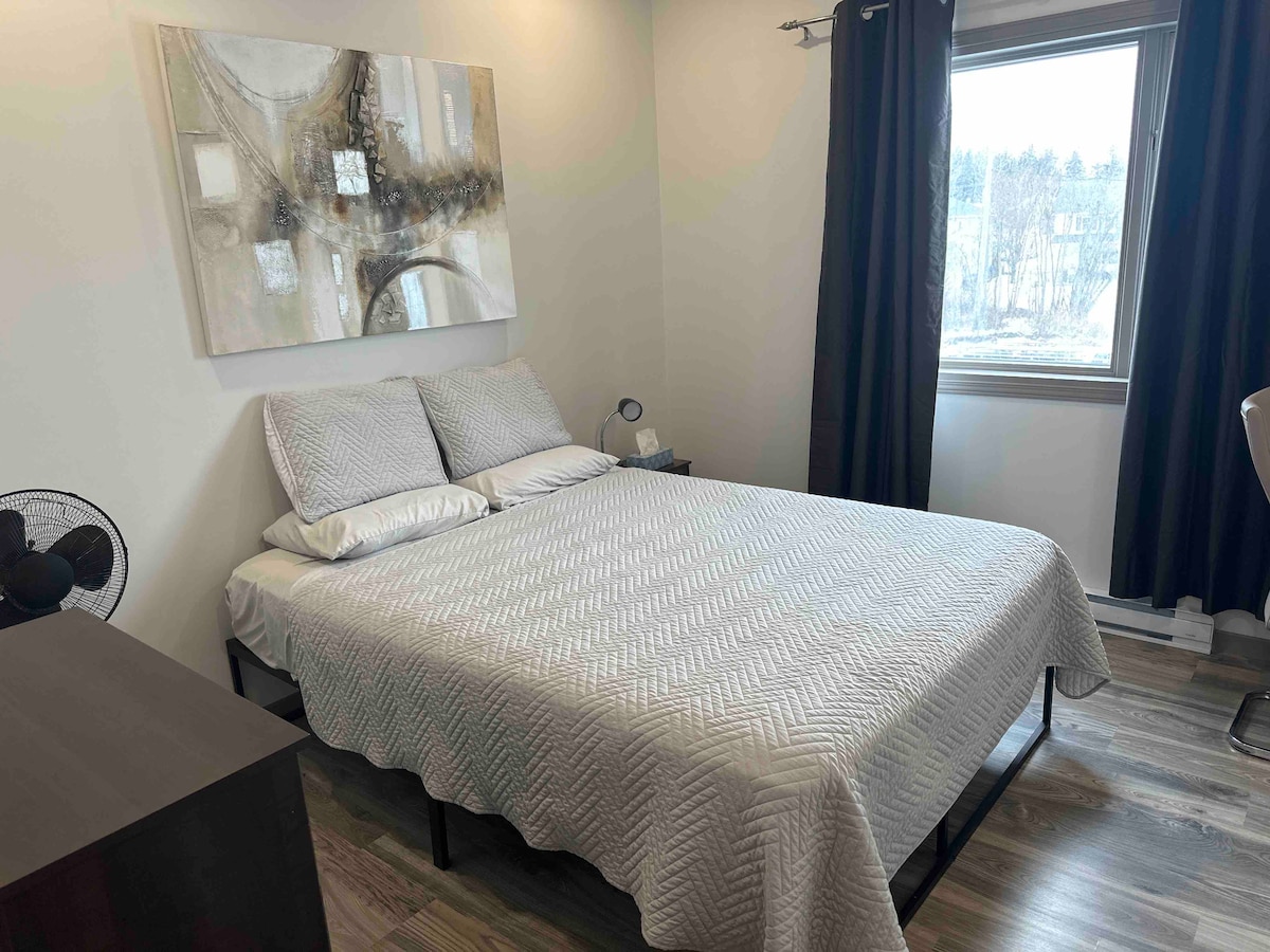 A well-lit bedroom is furnished with a spacious bed covered in a light quilt. The wall artwork complements the neutral tones, while a window dressed with dark curtains offers natural light. A nightstand and a small fan are positioned beside the bed.