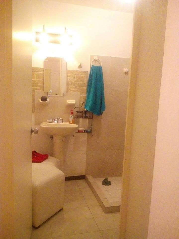A compact bathroom is visible, featuring a sink with a large mirror above and a shower area enclosed by a glass partition. A chair is positioned nearby, and a turquoise towel is hanging from a hook. The floor is tiled, and the space appears well-lit.