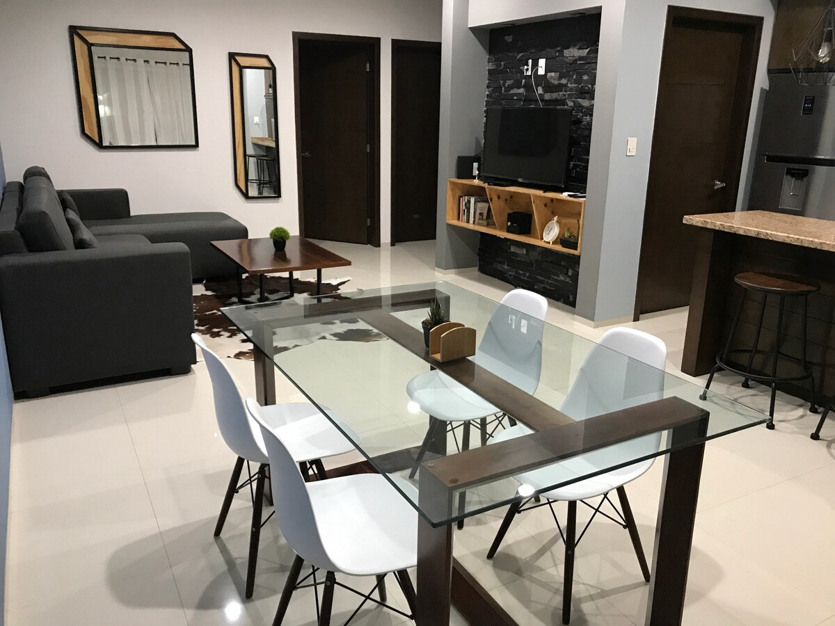 A modern living space features a glass dining table surrounded by four white chairs. A brown wooden coffee table is positioned in front of a dark grey sofa. A flat-screen TV is mounted on the wall, with decorative shelving nearby, enhancing the contemporary feel of the area.