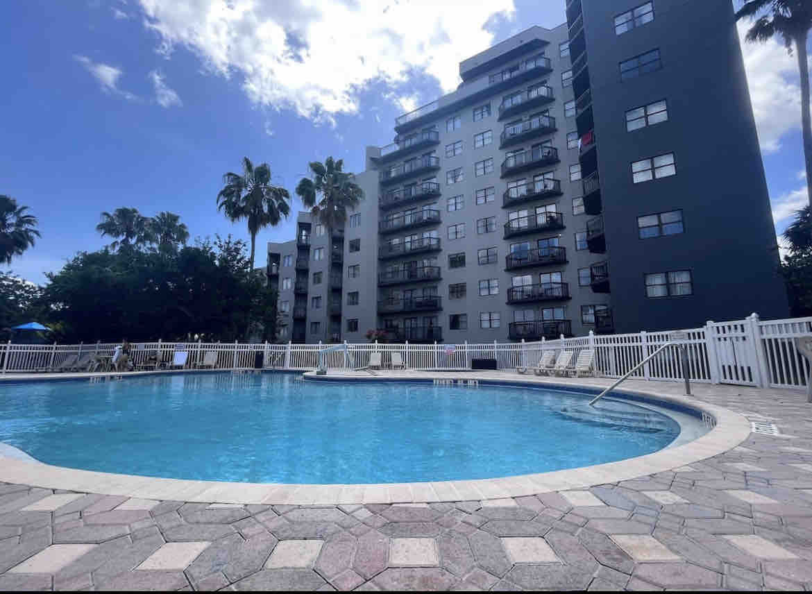 A spacious swimming pool is surrounded by a light-colored paved deck and a white fence. Several palm trees are visible in the background, along with a multi-story building featuring balconies. A clear blue sky enhances the inviting atmosphere of the pool area.