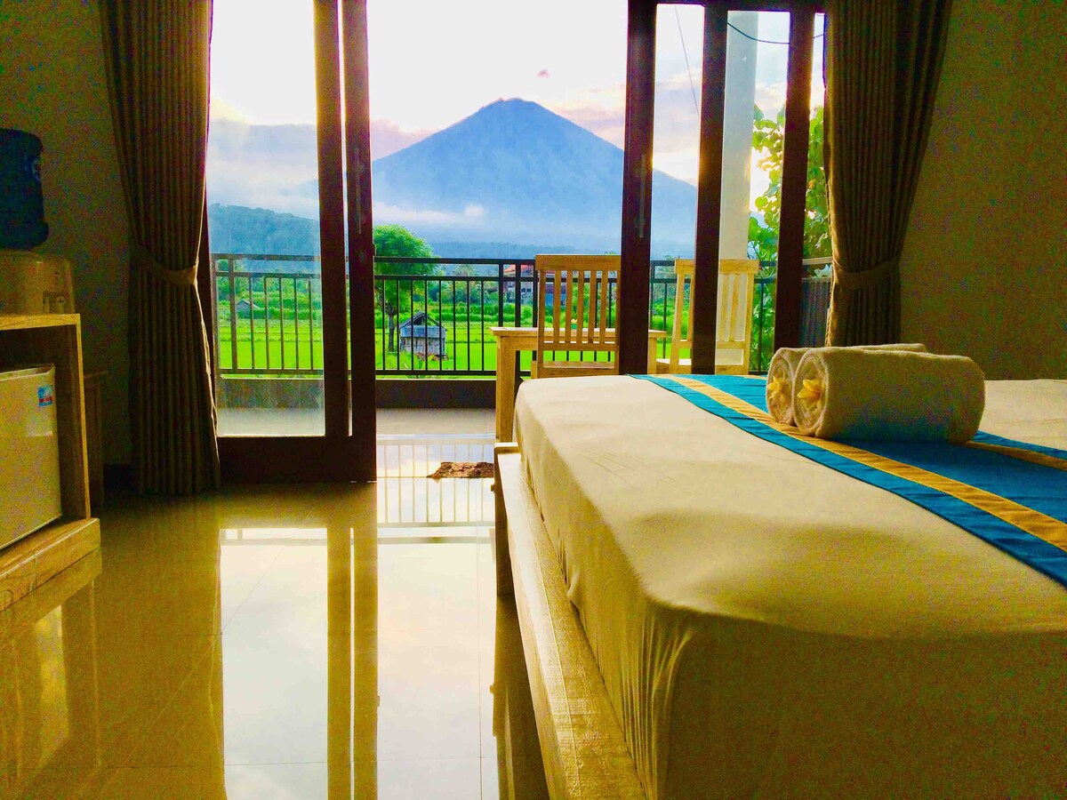 A spacious room features a large bed with a simple, neutral design. Large glass doors open to a balcony showcasing a panoramic view of Mount Agung and surrounding rice fields, while soft light fills the room from the sunset.