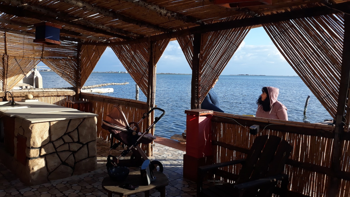 An open space features a rustic stone countertop surrounded by natural wooden structures. Large openings provide views of the calm water and distant shoreline, with a comfortable seating area visible. A bright atmosphere is created by daylight streaming through the woven canopies.