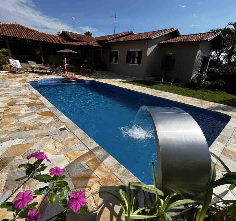 A private outdoor area features a blue swimming pool with a water fountain. Surrounding the pool are stone tiles and colorful flowers. A shaded seating area with umbrellas is visible, alongside the house, which showcases a tiled roof and several windows.