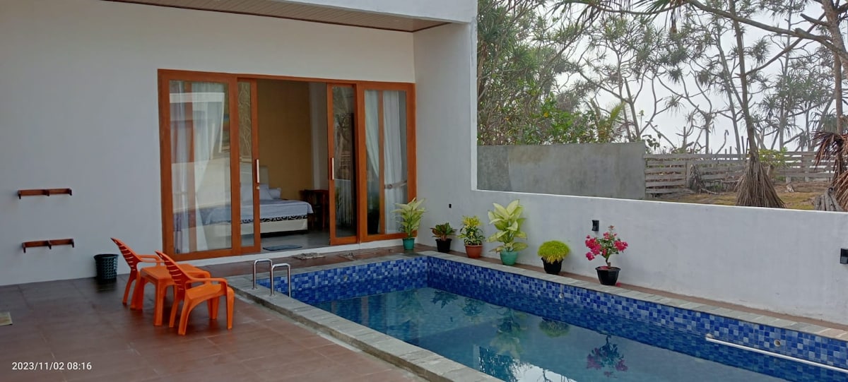 A private pool area is framed by light-colored walls and wooden sliding doors that open to an inviting interior. Potted plants are placed along the edge of the pool, enhancing the serene outdoor space. The tranquil water reflects surrounding greenery.