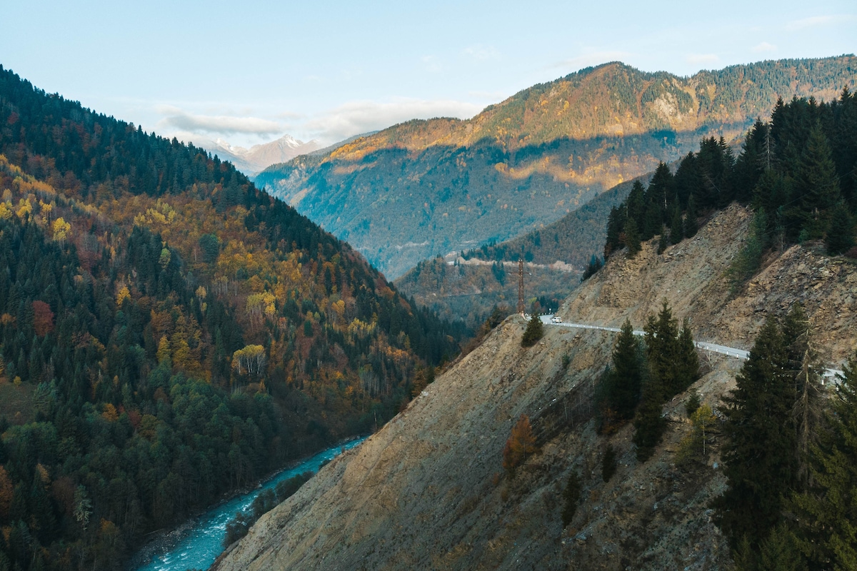 A panoramic view of mountainous terrain showcases autumn foliage on the slopes, with a winding road traversing the landscape. A river can be seen flowing through the valley below, surrounded by dense trees and rocky outcroppings under a clear blue sky.