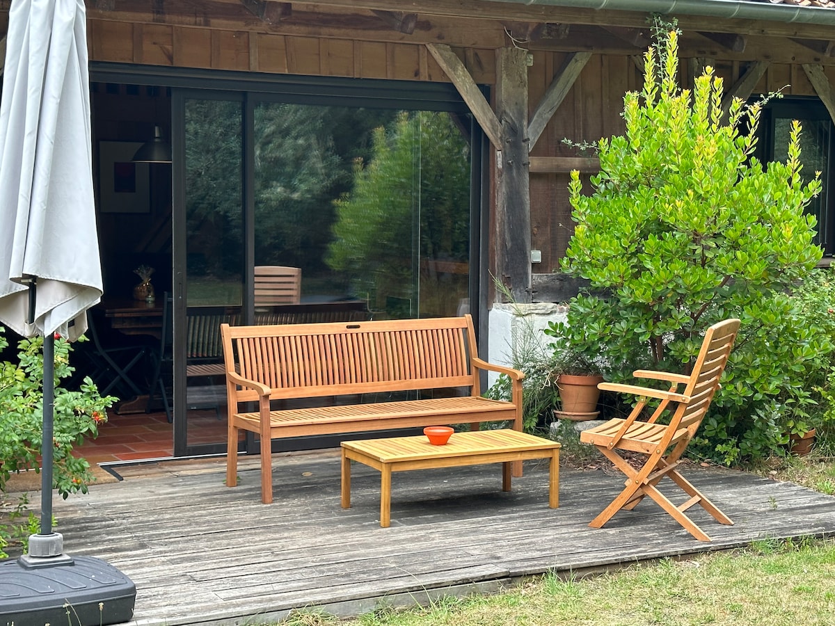 An outdoor seating area features wooden furniture, including a bench and a chair facing a coffee table. Glass doors provide a view into the interior space, surrounded by greenery that enhances the tranquil setting.