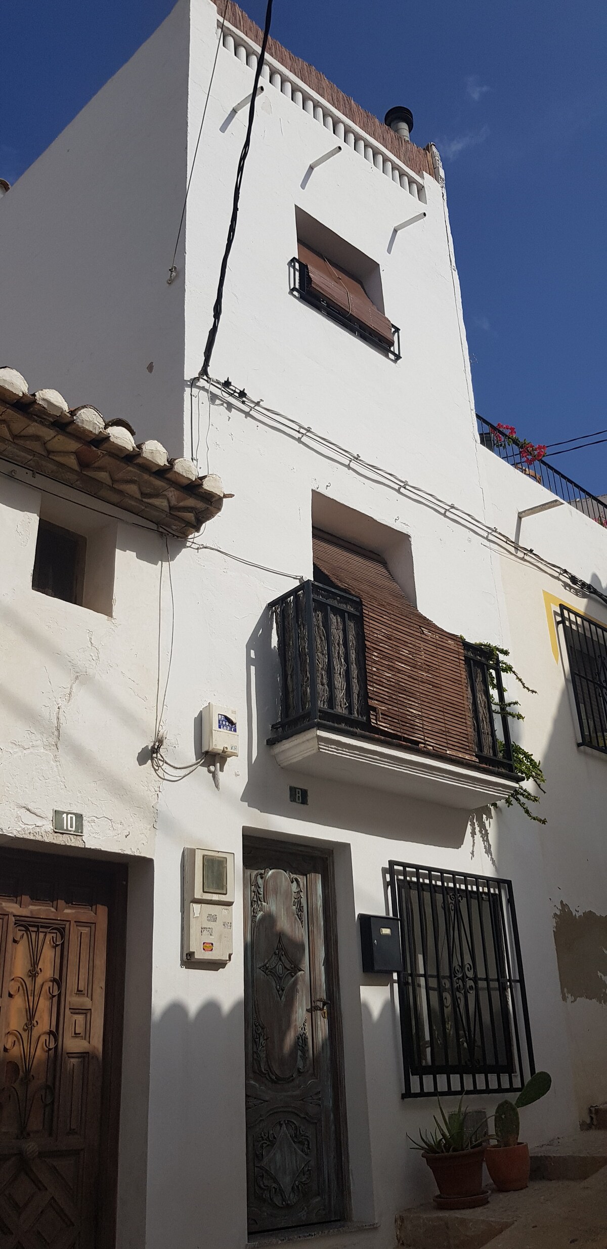 The exterior of a whitewashed townhouse is presented, featuring multiple levels and decorative wrought iron balconies. A rustic wooden door is visible at the entrance, surrounded by potted plants. The clear blue sky above adds to the charm of this quaint, village-style home.