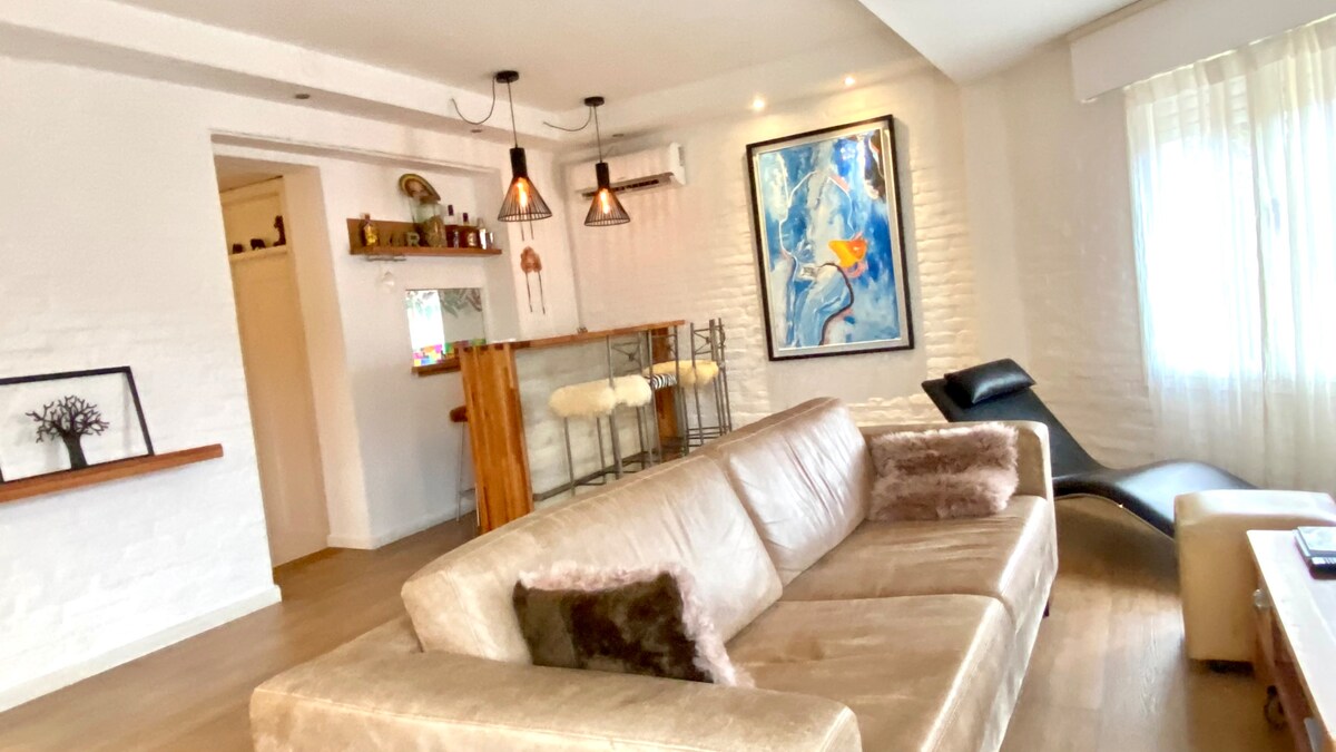A modern living area features a spacious, light-colored sofa with decorative cushions. A sleek bar with tall stools is visible in the back, accompanied by a large piece of wall art. Natural light is allowed in through sheer curtains, creating a bright ambiance.