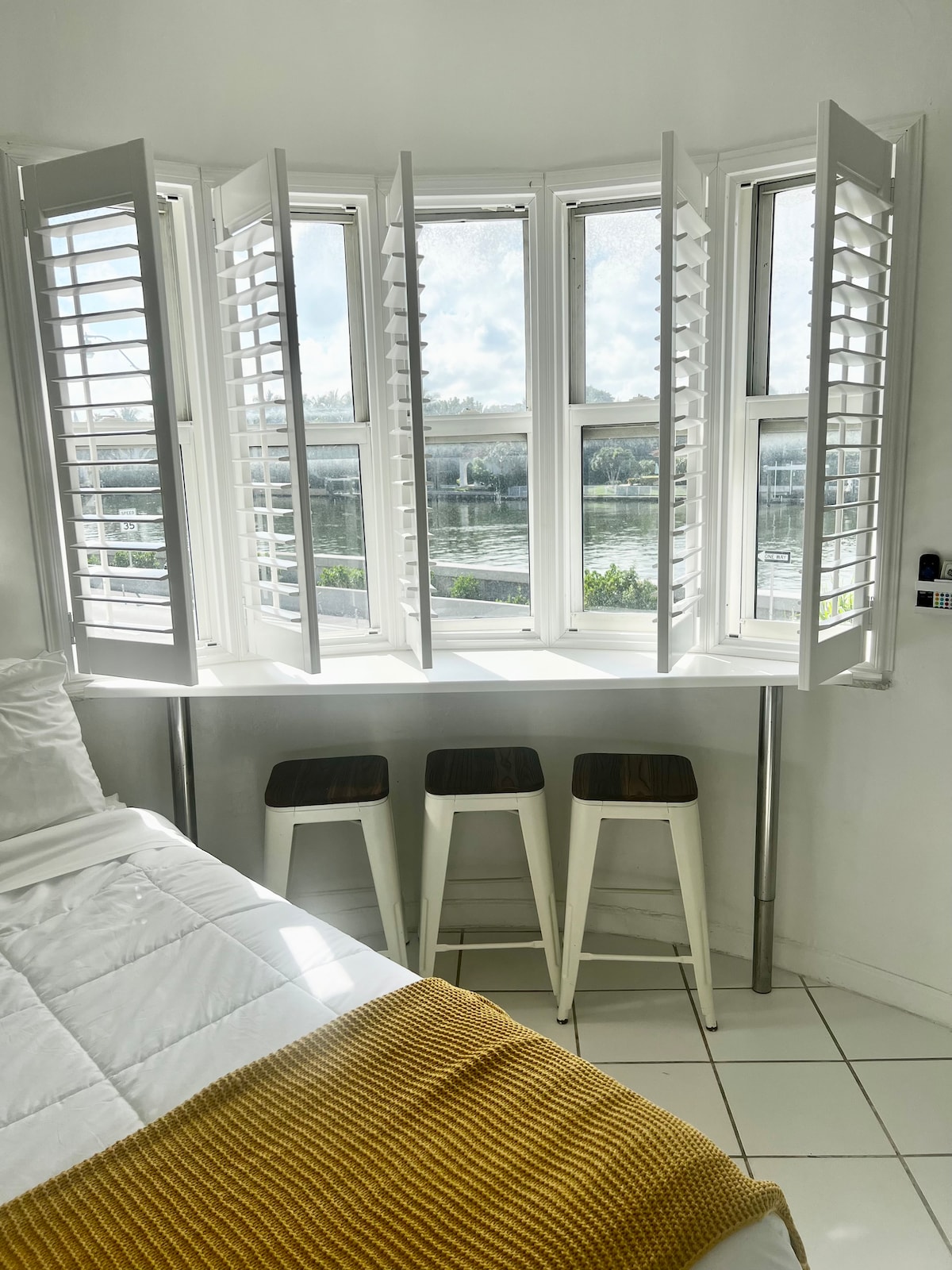 An inviting window nook features white shuttered windows that allow natural light to brighten the space. Below, three minimalist stools are positioned against a sleek countertop, providing a cozy spot for enjoying views of the water and surroundings.