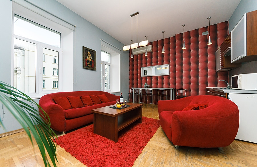 A spacious living area is furnished with red sofas and a matching rug. A wooden coffee table is positioned in the center. Behind the seating, a dining area with stools and a bar-style table is visible. Large windows allow natural light to enter, complementing the contemporary design.