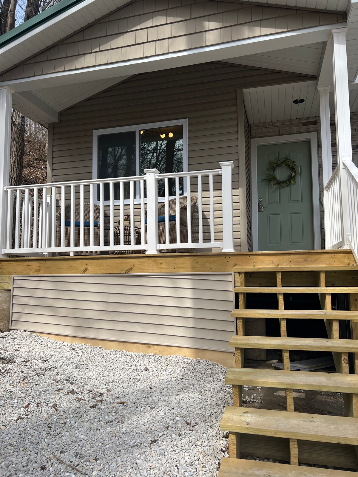 The exterior entrance features a welcoming porch area with white railings and steps leading up to the door. A light green door is framed by a small wreath, bringing a touch of charm to the cabin's exterior. Gravel surrounds the entrance, providing a natural setting.