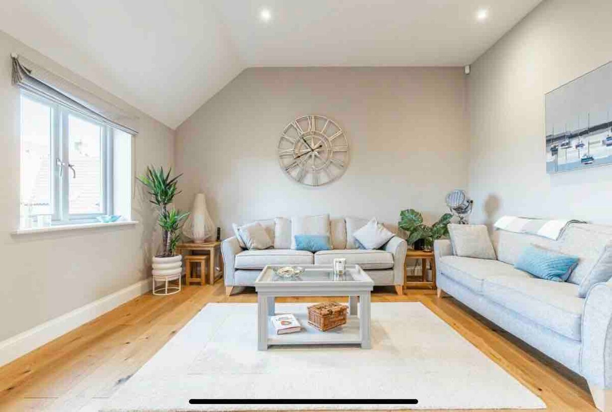 A bright living space is presented, featuring a light grey sofa and a matching loveseat, complemented by soft blue cushions. A large clock adorns the wall, while a decorative table sits on a neutral rug. Natural light streams through the window, enhancing the airy atmosphere.