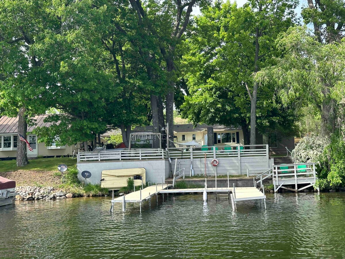A tranquil lakeside scene showcases a charming cottage surrounded by tall trees. A large wooden pier extends into the water, with several green chairs placed for relaxing. The patio area features seating, while a small dock is visible for convenient access to the lake.