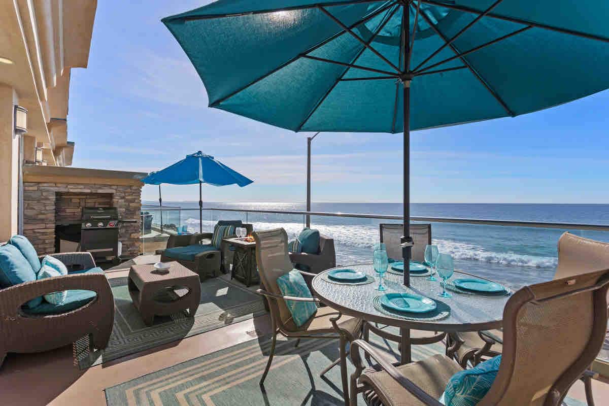 A private balcony features a dining table set for four, surrounded by comfortable seating areas. Vibrant blue umbrellas provide shade, while an ocean view stretches across the horizon. A decorative outdoor fireplace and a grill are present, creating an inviting space for relaxation.
