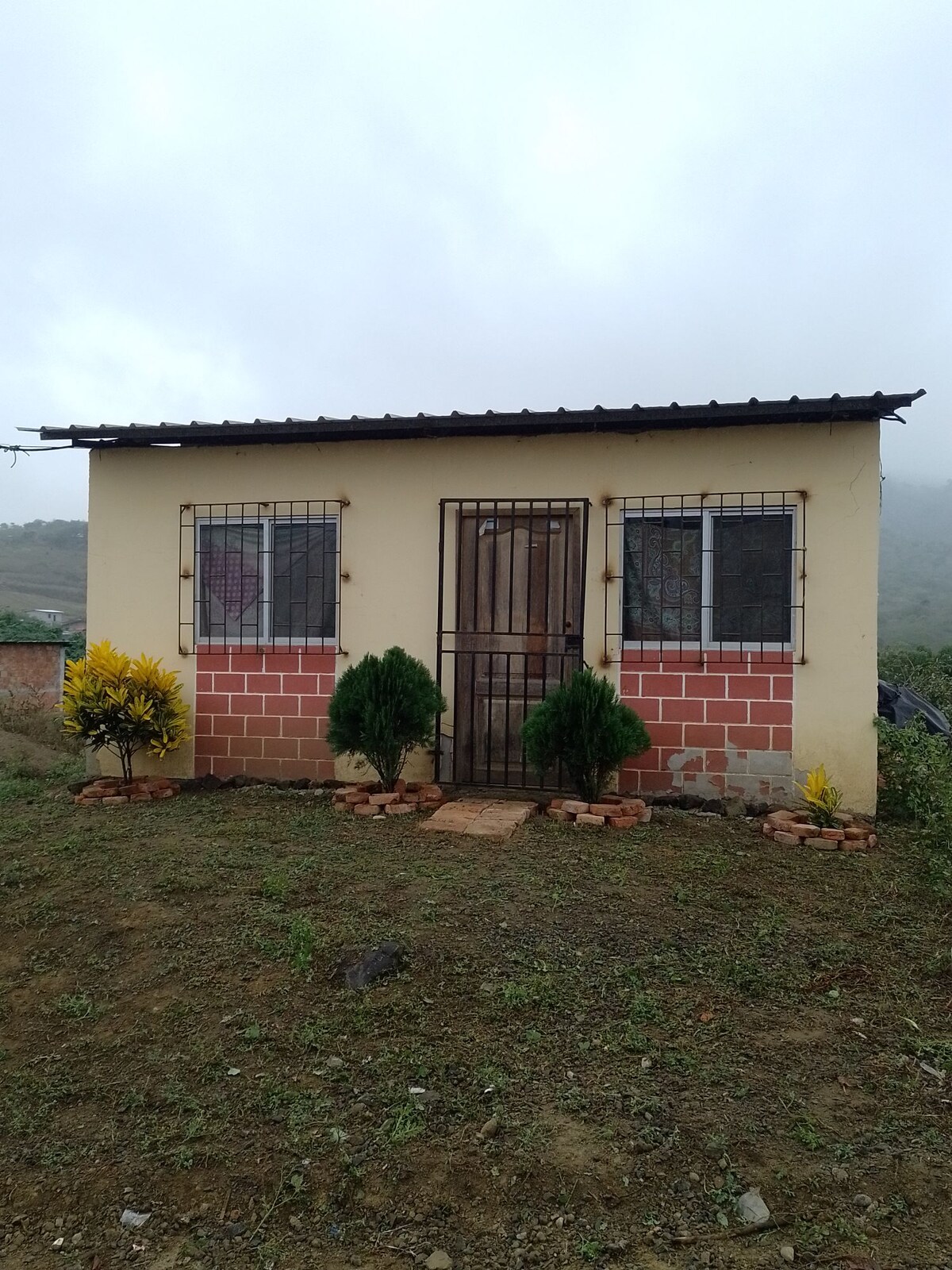 A small, charming house is shown with a simple facade featuring light yellow walls and a tiled roof. Two windows, secured by wrought iron grilles, are flanked by small, green shrubs. A stone path leads to a wooden door, enhancing the inviting exterior.
