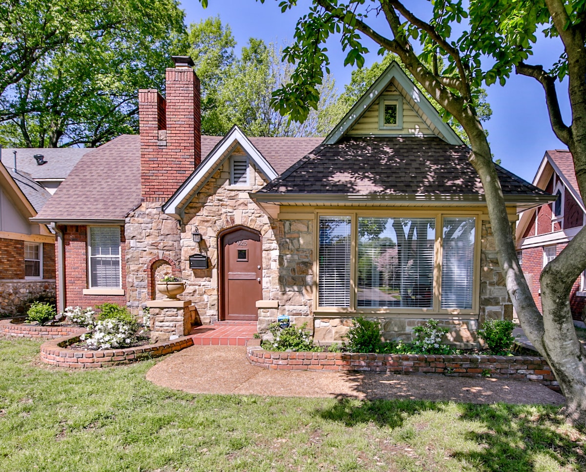 A charming stone exterior of a house is framed by green trees and a well-maintained lawn. The front entrance features a wooden door and large windows, allowing natural light to enter the interior. Flower beds with white blooms add a touch of color.