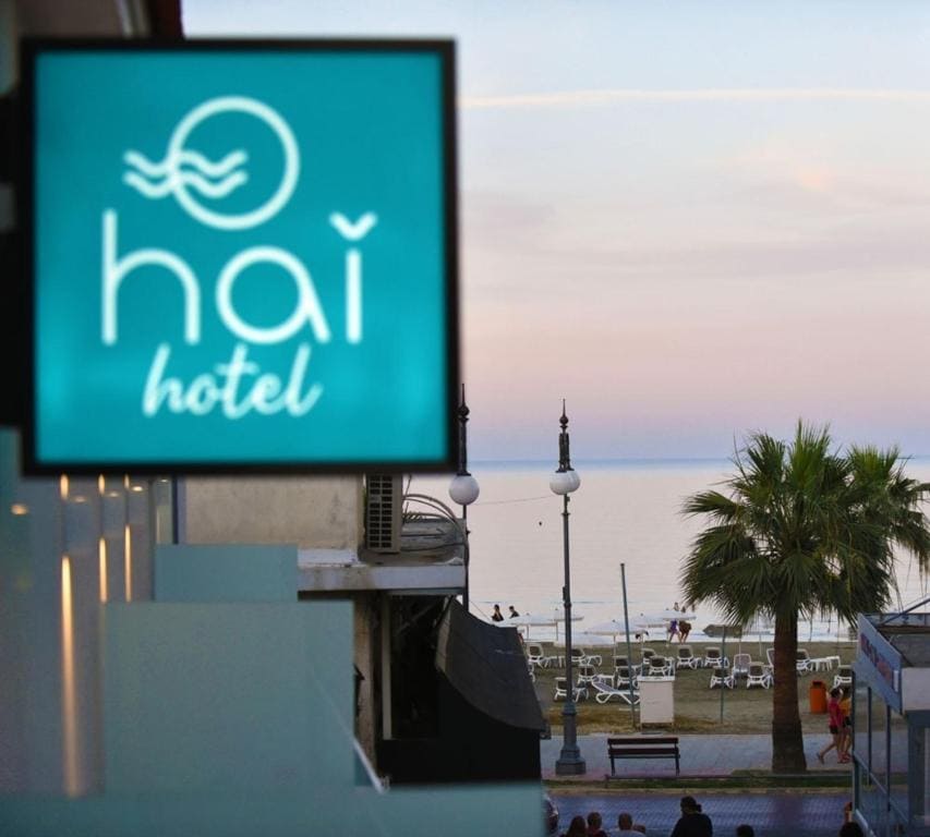 The image displays the Hai hotel sign against a backdrop of a calm seaside view. Palm trees and beach chairs are visible along the shoreline, under a soft sunset sky that gradually transitions from pink to blue.