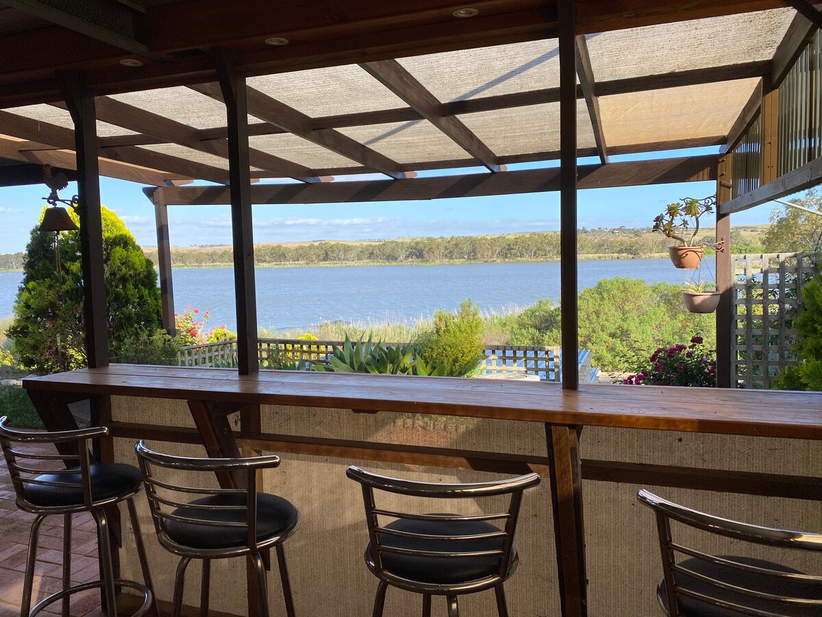 A shaded outdoor bar area provides seating with metal chairs facing the river view. A landscape of greenery and colorful flowers is visible in the background. The water reflects the blue sky, creating a serene atmosphere.