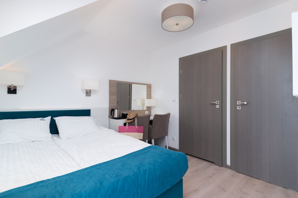 A well-appointed room features a spacious bed with crisp linens, complemented by a plush teal headboard. A modern desk and chair are positioned nearby, along with a mirror and a small table. Two light fixtures provide soft illumination, and two doors lead to adjoining spaces.