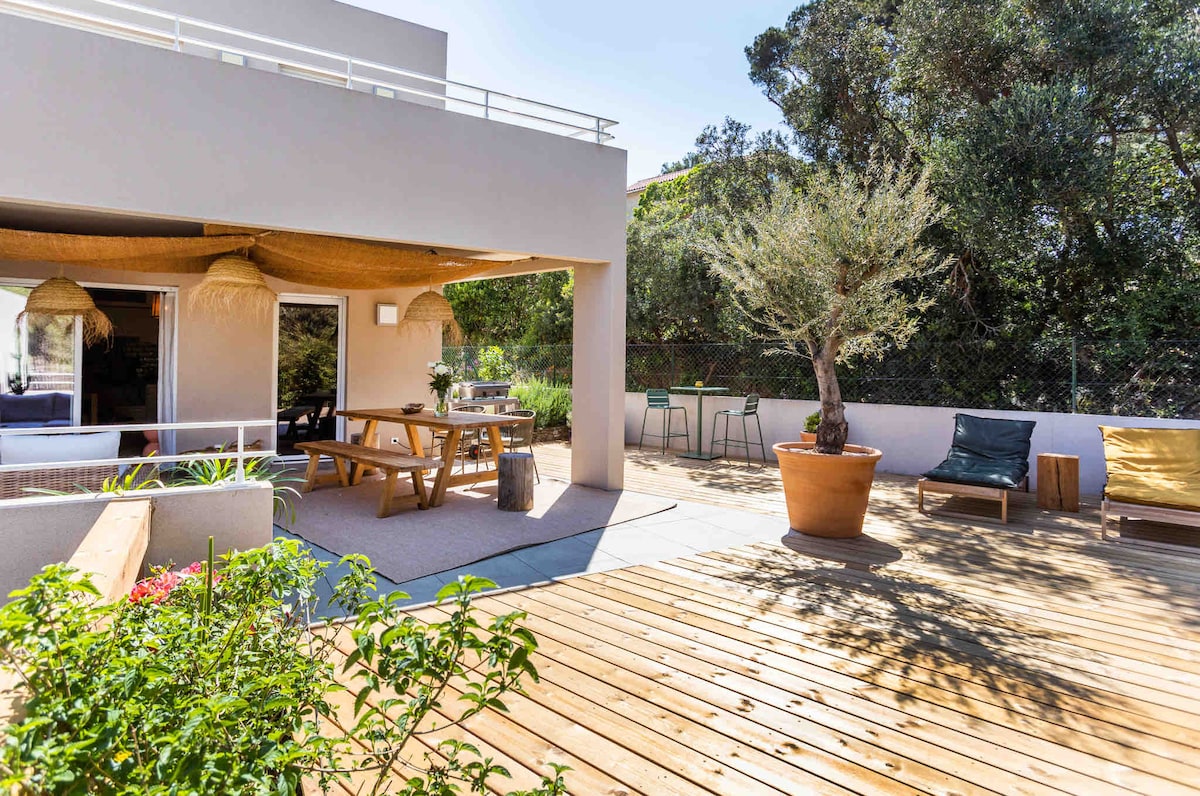 An outdoor terrace area showcases wooden decking and a spacious seating arrangement. Potted plants and greenery surround the space, which includes a dining table and chairs. Natural light enhances the ambiance of the area, providing a comfortable setting for relaxation.