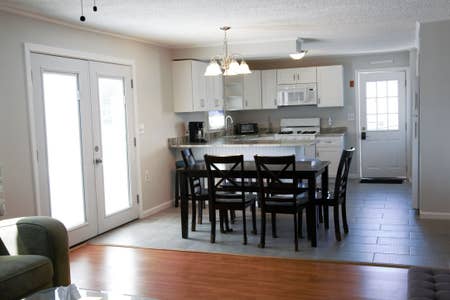 An open kitchen and dining area are visible, featuring a modern kitchen with white cabinets, a gas stove, and granite counters. A dining table with four black chairs is positioned nearby. Light enters through double doors, enhancing the clean, neutral décor.