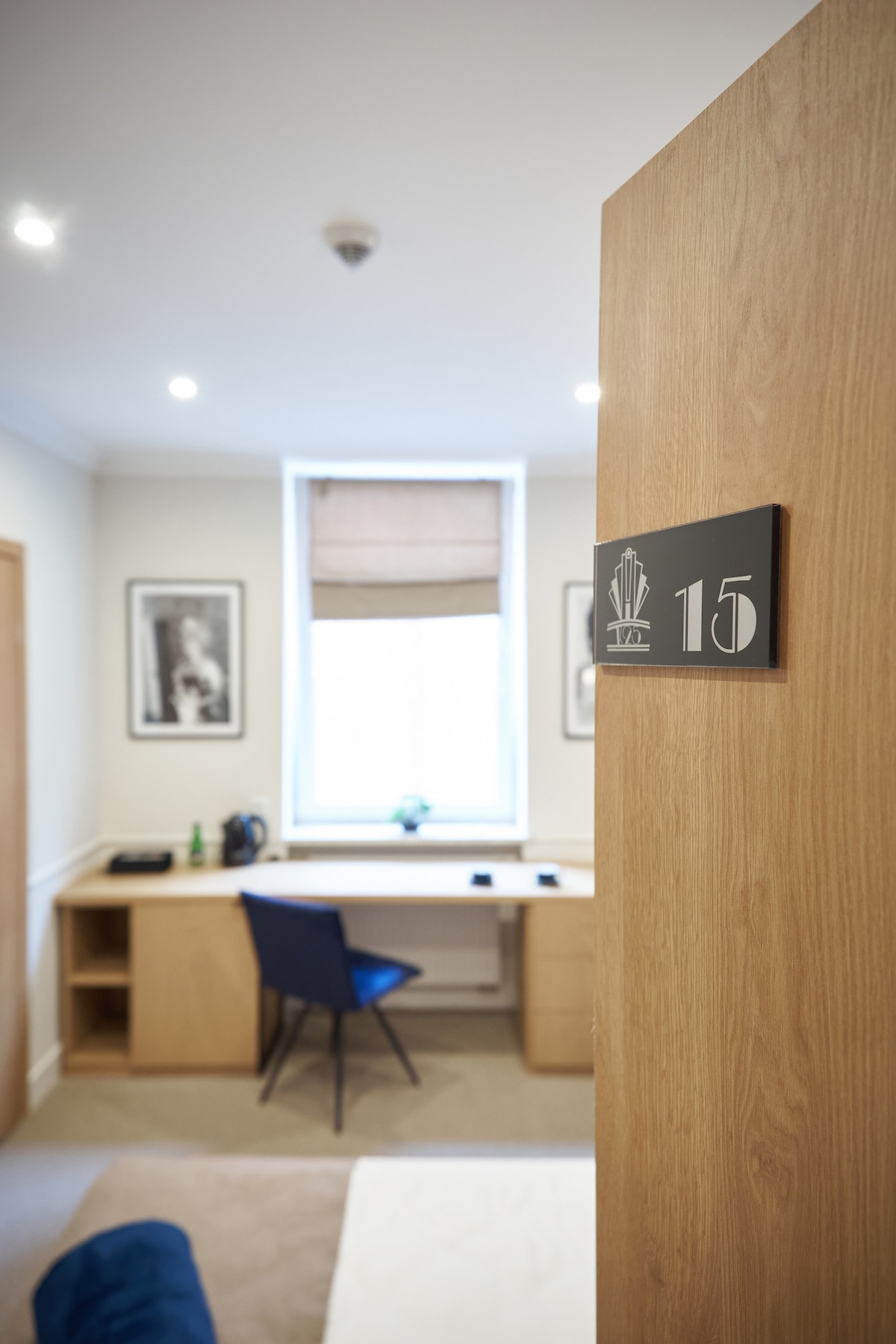 An inviting room is accessed through a wooden door, featuring a modern desk with a chair and decorative artwork on the walls. Natural light enters through a window with a light-dimming shade. A small plant adds a touch of nature to the workspace.