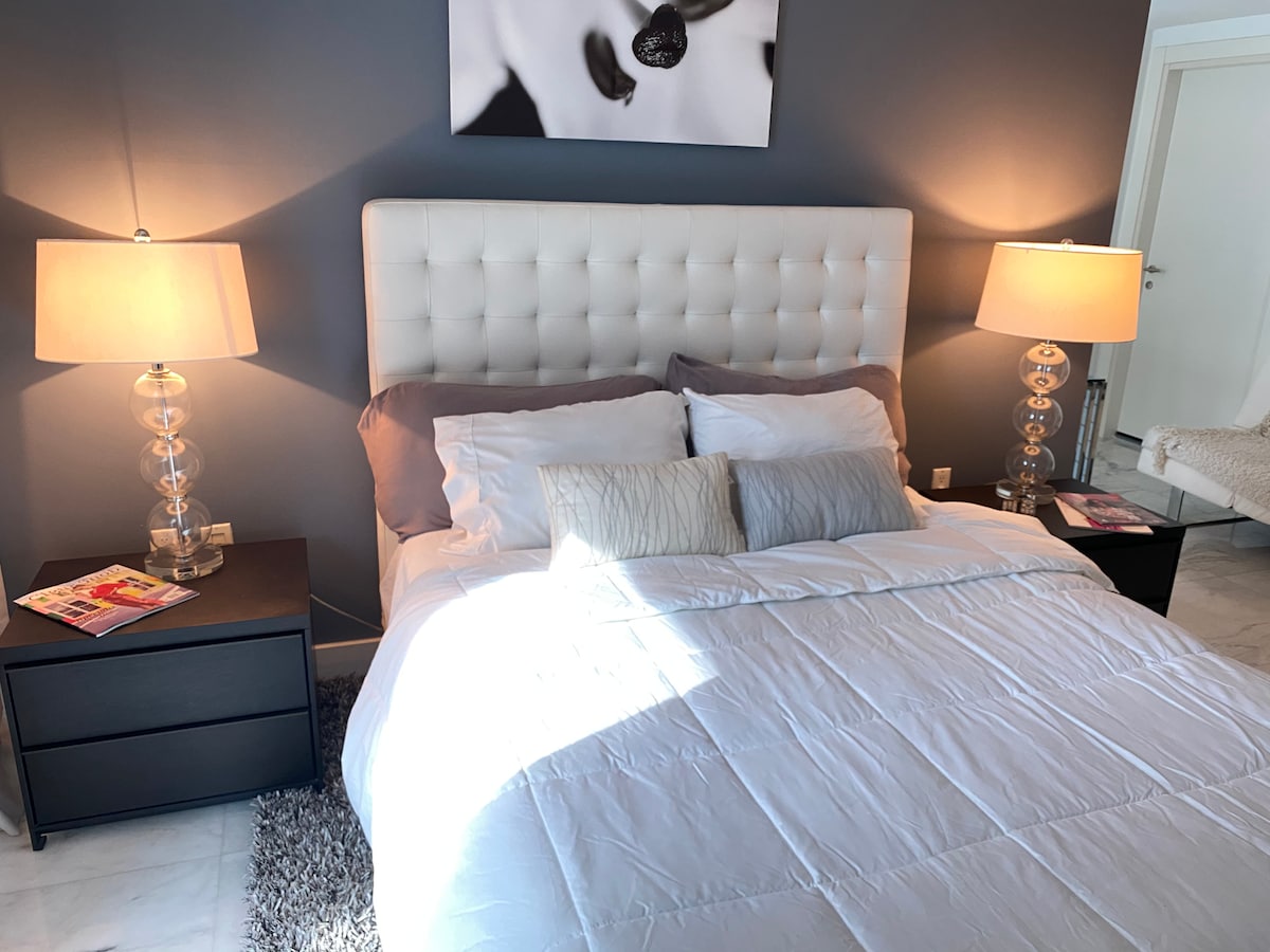 A bed with a tufted headboard is centered against a dark wall, complemented by two bedside tables with decorative lamps. Soft sunlight filters into the room, highlighting the white bedding and pillows. Magazines are placed on the tables, adding a personal touch.
